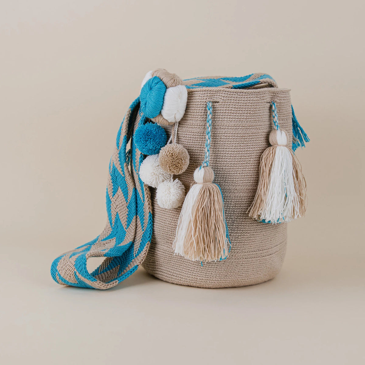 Front view of Cartagena Wayuu Mochila featuring beige handwoven design, turquoise accents, decorative pom-poms and tassels – Maison Monserrate